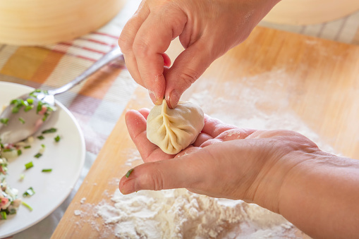 Female hands are preparing manti, dumplings or wonton from dough with meat filling on the background of a kitchen table with flour. Close-up.