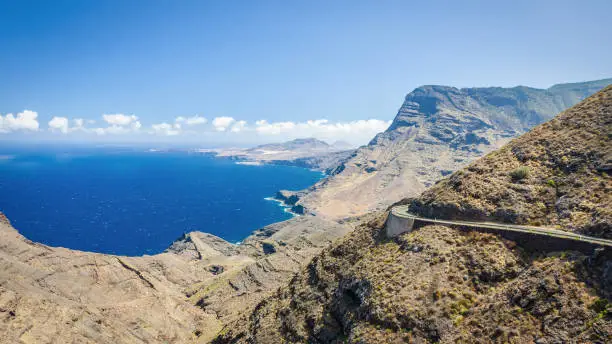 Panorama view over the rocky coast of Gran Canaria in Tamadaba Natural Park with winding road through dry rocky coastal mountain landscape under blue summer sky. Tamadaba Natural Park, Grand Canary, Canary Islands, Spain.