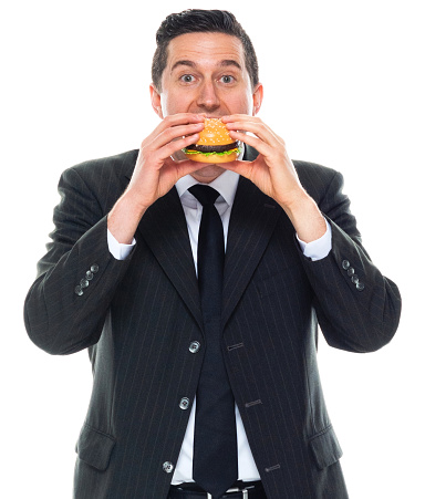 Portrait of caucasian male manager standing in front of white background wearing businesswear who is hungry and holding cheeseburger