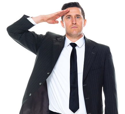 Front view of caucasian male business person standing in front of white background wearing businesswear who is smiling and showing hand raised who is saluting with arms raised