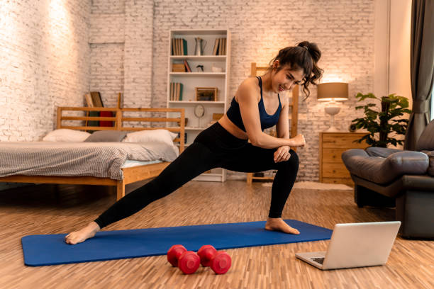 20s young Asian woman in sportswear doing stretching exercises while watching fitness training class on computer laptop online. Healthy girl exercising in living room with sofa couch and bed in the background.. mujer joven asiática de los 20 años en ropa deportiva haciendo ejercicios de estiramiento mientras ve la clase de entrenamiento de yoga en computadora portátil en línea. chica sana haciendo ejercicio en la sala de estar con sofá sofá en el fondo.