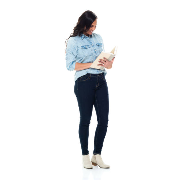 Caucasian female student standing in front of white background wearing jeans and holding book - fotografia de stock