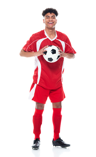 Full length of aged 18-19 years old with curly hair african ethnicity teenage boys athlete standing in front of white background wearing soccer uniform who is laughing and holding soccer ball and playing soccer - sport and using sports ball