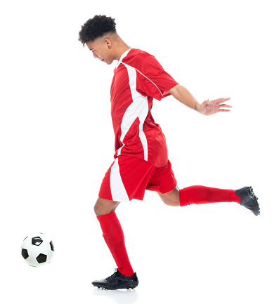 Side view of aged 18-19 years old with curly hair generation z male soccer player jogging in front of white background wearing soccer uniform who is in concentration who is fighting and holding soccer ball and playing soccer - sport and using sports ball