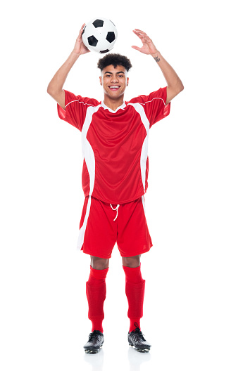 Full length of aged 18-19 years old with black hair african-american ethnicity young male athlete standing in front of white background wearing soccer uniform who is smiling and holding soccer ball and playing soccer - sport and using sports ball