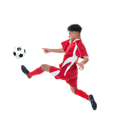 Full length of aged 20-29 years old with black hair african-american ethnicity young male soccer player mid-air in front of white background wearing soccer uniform who is serious and holding soccer ball and playing soccer - sport and using sports ball