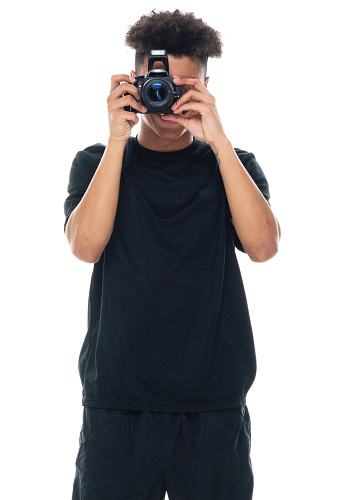 Front view of aged 18-19 years old with curly hair african-american ethnicity young male photographer standing in front of white background wearing sports shoe who is smiling who is photographing and holding camera