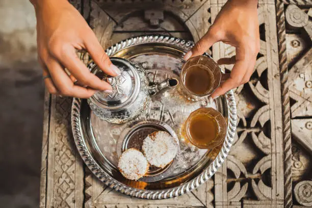 Photo of Woman hands serving traditional moroccan mint tea ceremony with cookies and vintage silver teapot. Hospitality and service in Morocco, Marrakech.