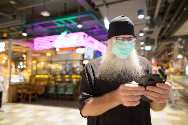istock Mature bearded hipster man using phone with mask for protection from corona virus outbreak at the mall 1224102536