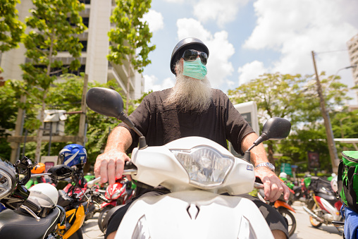 Portrait of mature bearded hipster man with mask for protection from corona virus outbreak riding motorcycle in the city outdoors