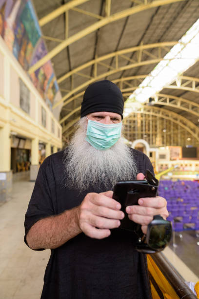 istock Mature bearded tourist man using phone with mask for protection from corona virus outbreak at railway station 1224102770