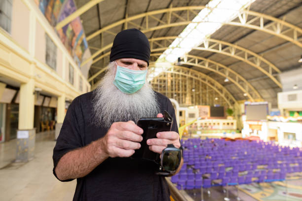 istock Mature bearded tourist man using phone with mask for protection from corona virus outbreak at railway station 1224102776