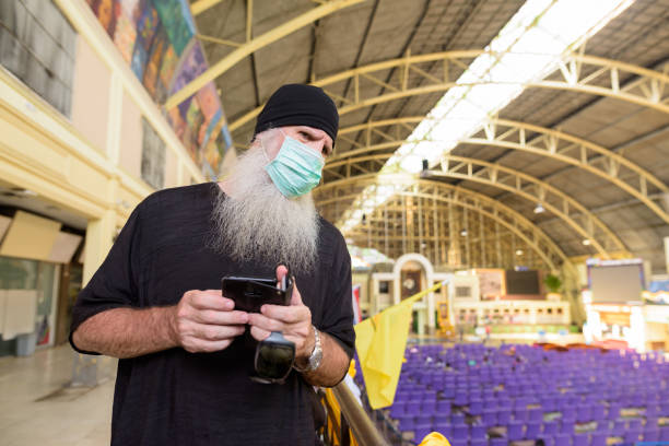istock Mature bearded tourist man with mask thinking while using phone at railway station 1224102777