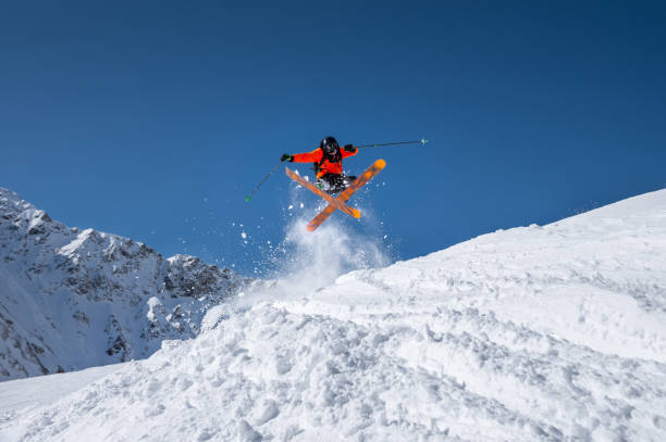 esquiador atleta masculino em um gatilho laranja faz um truque de salto com pó de neve voadora contra o pano de fundo das montanhas do cáucaso cobertas de neve e um céu azul. conceito de esportes radicais de inverno - skiing winter sport powder snow athlete - fotografias e filmes do acervo