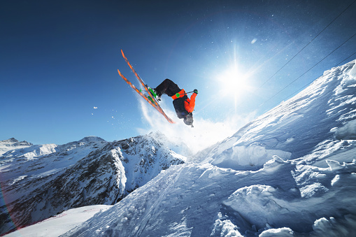 Low angle view athlete skier in an orange jacket does a back flip with flying powder of snow against a clear blue sky sun and snow-capped mountains of the Caucasus. Winter Extreme Sports Concept