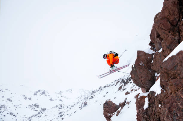 曇りの日に雲の中の山々を背景に、高い岩の雪の斜面から飛び降りる灰色のバックパックとスキーポールを持つオレンジ色のトリガーで男性アスリートスキーヤー。 - powder snow skiing agility jumping ストックフォトと画像