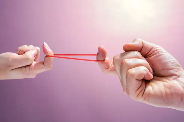 Close-up of man and woman stretching red rubber band with fingers against pink background.