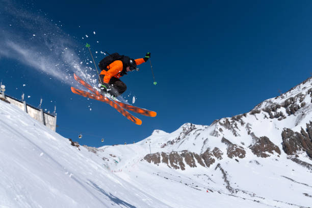 um esquiador atleta masculino em um gatilho laranja faz um salto com um agarramento com pó de neve voadora contra o fundo do monte elbrus no cáucaso norte. conceito de esportes radicais de inverno - skiing winter sport powder snow athlete - fotografias e filmes do acervo