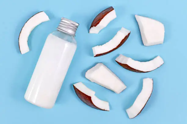 Photo of Coconut oil bottle used for food preperation and cosmetic beauty products surrounded by slices of coconut fruit on blue background