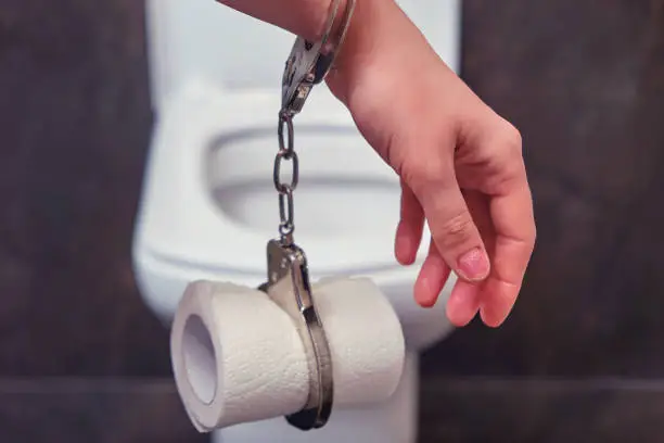 Photo of Hand chained by handcuffs to toilet paper on the background of the bowl, close-up. The concept of stomach disease, poisoning and diarrhea.