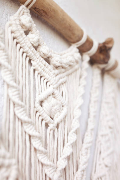 istock Handmade macrame  100% cotton wall decoration with wooden stick hanging on a white wall.  Macrame braiding and cotton threads. Details close up. Female hobby. 1224125321