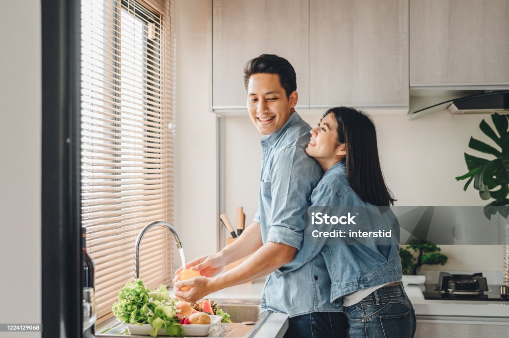Asian couple lovers hug in the kitchen while cooking at home Asian couple lovers hug in the kitchen while cooking at home. A woman hugs a man who is washing vegetables from the back. Couple - Relationship Stock Photo