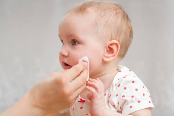 若い母親の手は白い綿パッドで女の赤ちゃんの頬を拭きます。日常。幼児の清潔で柔らかい体の皮膚を気にします。クローズ アップ。サイドビュー。 - human face rubbing women beauty treatment ストックフォトと画像