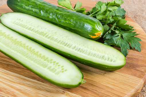 One whole and two halves of long green smooth-skinned cucumbers on the wooden cutting board with fresh parsley close-up in selective focus