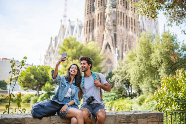 Young Couple Taking Break from Sightseeing for Selfie Male and female travelers sitting on wall in public park near Sagrada Familia in Barcelona and taking selfie on sunny summer day. photo messaging stock pictures, royalty-free photos & images