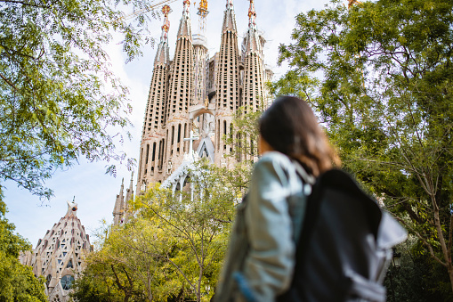 Low angle view with focus on background of young Indian female backpacker walking past camera through public park on the way to Sagrada Familia.