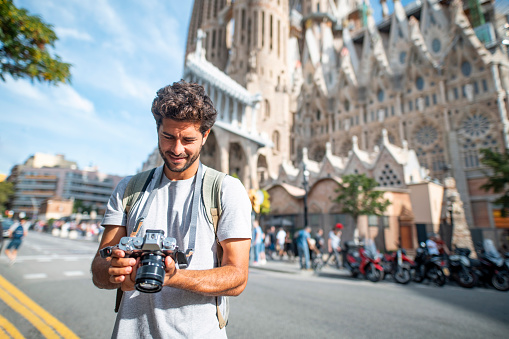 Front view of male tourist in early 30s using camera while sightseeing in sunny Barcelona with Sagrada Familia in background.
