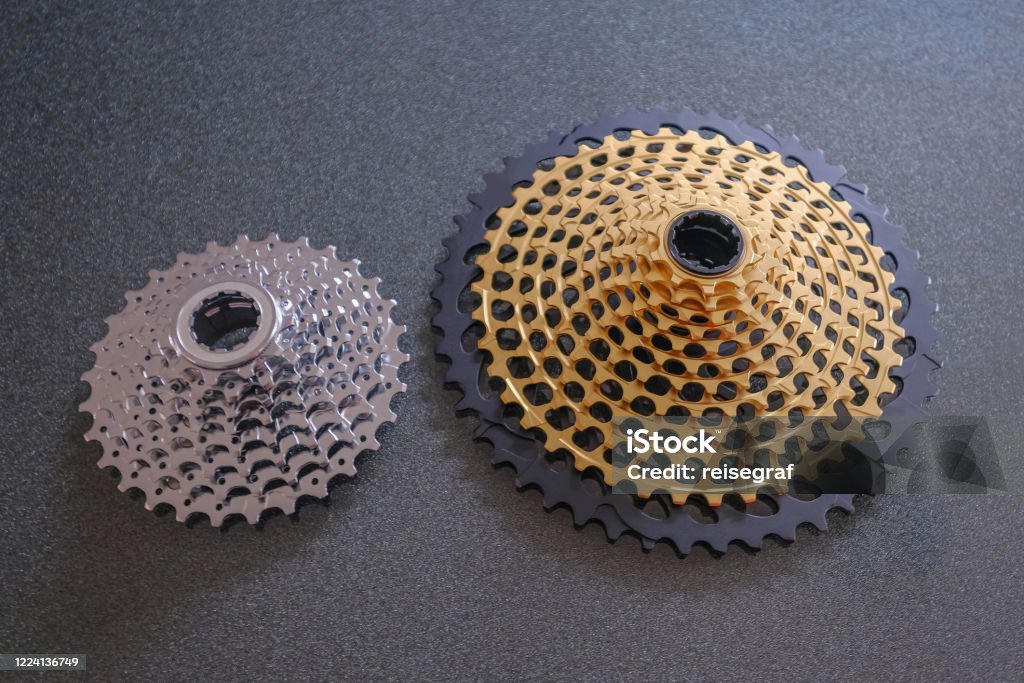 Rear mountain bike cassette, size comparison 12-speed vs 8-speed, small and big, in silver and golden color Rear mountain bike cassette, size comparison 12-speed vs 8-speed, big and small, in silver and golden color, the big 12-speed one is a 10-50 teeth Bicycle Chain Stock Photo