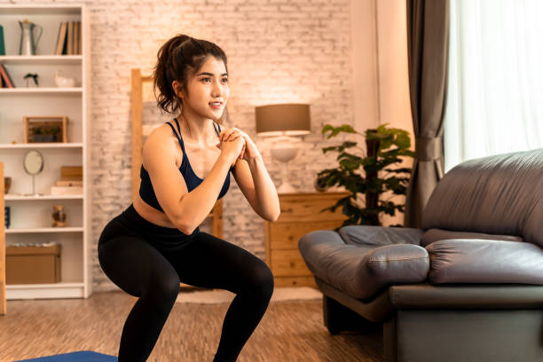 Young fit Asian woman working out at home. Beautiful female athlete training for legs muscles with squats exercise move Young fit Asian woman working out at home. Beautiful female athlete training for legs muscles with squats exercise move. squatting position photos stock pictures, royalty-free photos & images