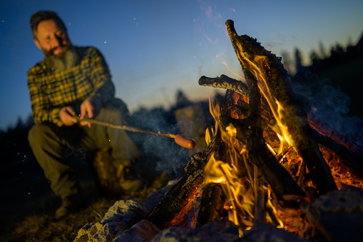 Mature man roasting sausage over campfire at dusk.