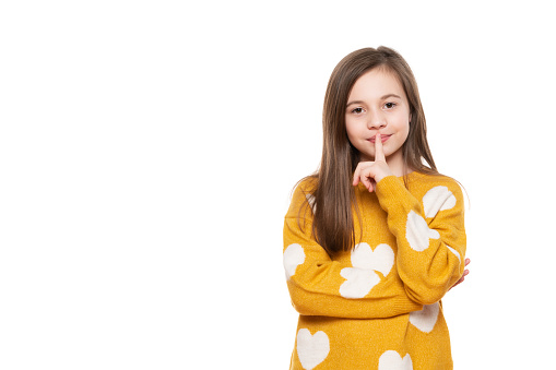 Finger on lips, silent gesture. Shhh! Waist up portrait of a charming young schoolgirl showing silence symbol, holding forefinger on lips, looking at camera, isolated on white background.