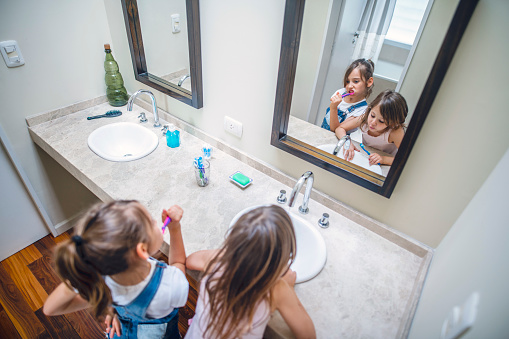 Elevated close-up of 6 and 8 year old sisters standing together at bathroom sink and brushing their teeth.