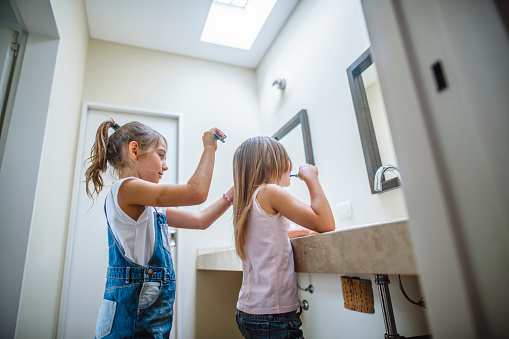 Close-up of younger sister brushing her teeth while older sister brushes her hair in family bathroom.