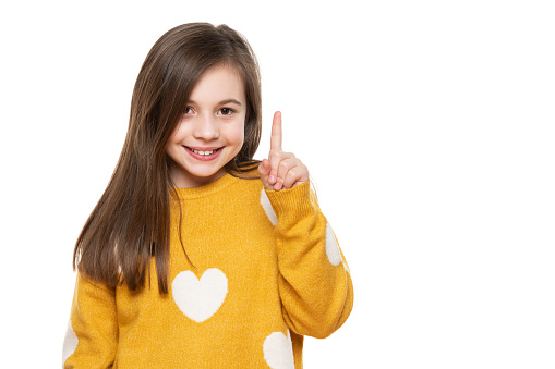 Beautiful young girl in mustard yellow sweater looking at camera, smiling and pointing up. Waist up studio shot on white background.