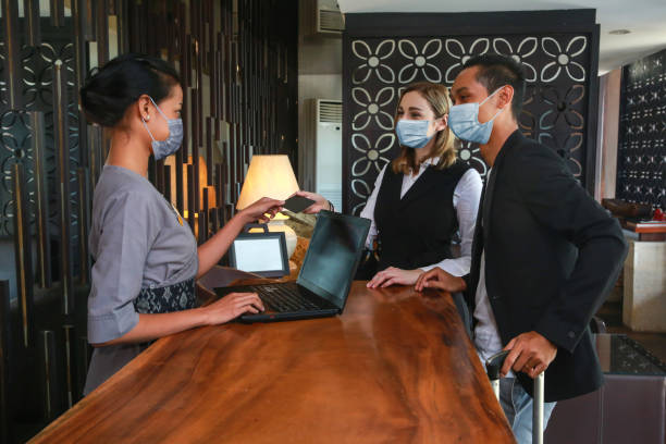 바이러스 예방 조치로 의료 용 마스크를 착용하고 있는 호텔 카운터의 커플 및 접수. 호텔에서 체크인을하는 출장에 커플 - 호텔 뉴스 사진 이미지