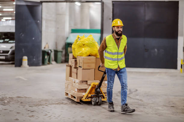istock Full length of smiling hardworking bearded worker in vest, with safety helmet on head pulling a pallet truck with boxes, sack and building material. Building in construction process interior. 1224171554