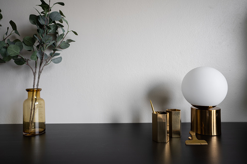 Stylish working corner decorated with mirror hexagon stainless vases, tint brown glass vase with artificial plant inside and gold mirror circular table lamp setting on black wood table top and beige color background /  copy space interior decoration