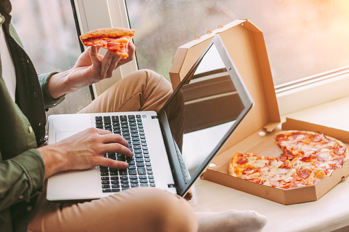 Closeup stylish businessman freelancer eat slice of tasty pizza and using laptop for distance work from home. Hipster man hold takeaway italian fast food snack in hand, type on keyboard. Lunch break