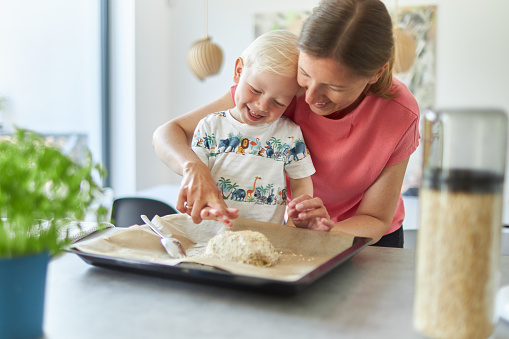 A mother bakes bread with her little blond-haired son in her modern kitchen and teaches him how to operate the food processor and wash his hands properly after having finished the dough. They laugh a lot together and you can see that they love each other very much.