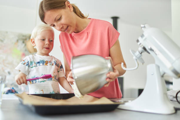 une mère fait cuire du pain avec son petit fils aux cheveux blonds dans sa cuisine moderne et lui apprend à faire fonctionner le robot culinaire et se laver les mains correctement après avoir fini la pâte. - wash bowl photos et images de collection