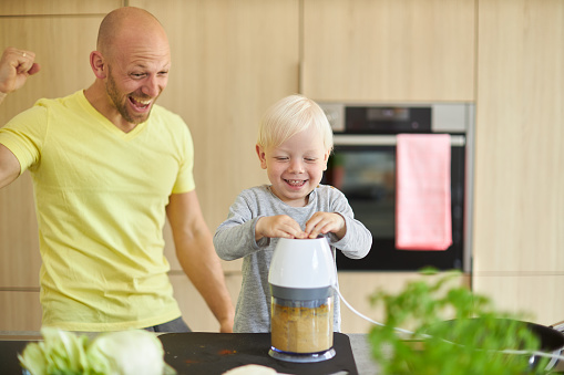 A father prepares a sandwich and homemade hummus with his son in the modern kitchen at home while he shows him and explains how to cook, cut vegetables and operate the blender and the garlic press. he wears a yellow t-shirt and have fun