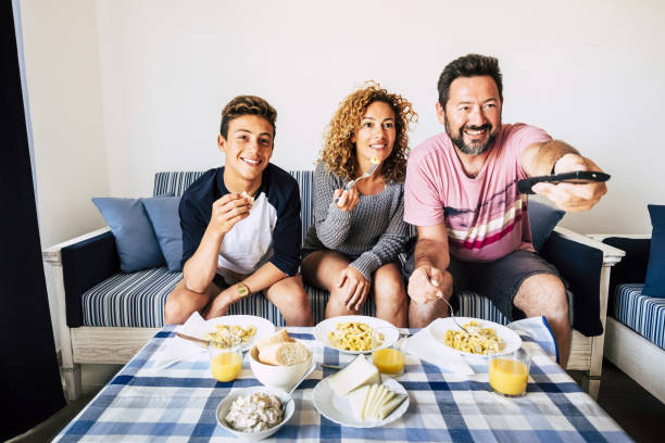 felici persone allegre della famiglia caucasica pranzano insieme a casa guardando la tv e divertendosi - il giovane figlio adolescente e i padri adulti sorridono e si godono l'attività indoor mangiando cibo - italian culture women looking at camera cheerful foto e immagini stock