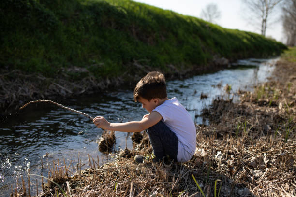 A young boy is squatting  on the edge of a creek and holding a stick as a fishing rod