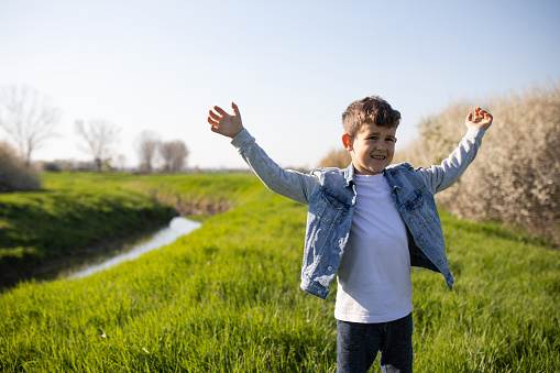 A cute boy is standing in tall grass next to a creek in the countryside in spring and spreading his arms wide