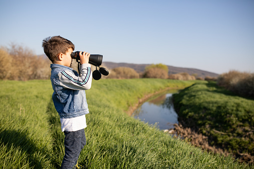A boy is standing next to a creek in the countryside looking through binoculars to see water birds in sprig