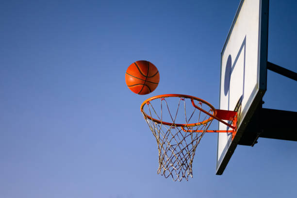 street-basketball-ball fällt in den reifen. nahaufnahme der orangen kugel über dem reifennetz mit blauem himmel im hintergrund. erfolgskonzept, punkteundinn und gewinnen. kopierraum - basketballkorb stock-fotos und bilder